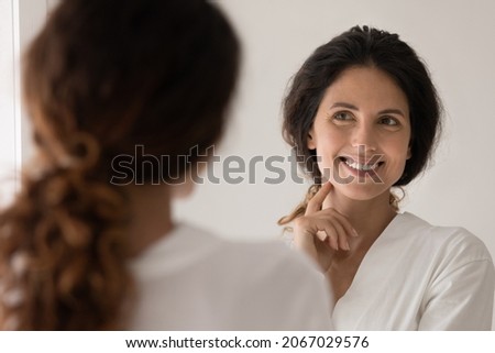 Beautiful Hispanic woman wear white bathrobe admire face view looking in mirror while standing in bathroom enjoy facial perfectness after skin procedure, applied cream, treatment for skincare beauty Royalty-Free Stock Photo #2067029576