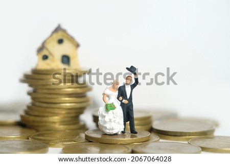 Housing for newlywed concept miniature people, toys photography. Bride and groom standing above coin pile in front of mini house isolated on white background. Image photo