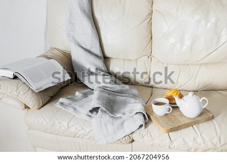 Cozy home cocnept. Teapot, cup of tea  and bun on the wooden try,open book, cozy blancket  on beige sofa for relax time at home. Large image for banner.