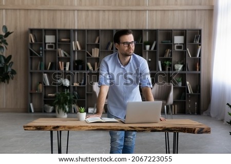 Pensive happy young businessman manager in eyeglasses looking in distance, visualizing future, thinking of challenges or opportunities standing at table with computer in modern workplace home office.