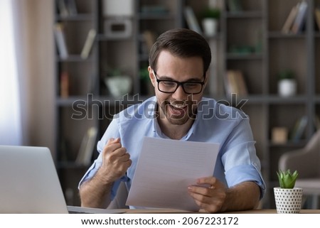 Overjoyed sincere laughing young man in eyeglasses making yes gesture, reading amazing news in paper correspondence, getting dream job offer, bank loan approval or celebrating signed contract. Royalty-Free Stock Photo #2067223172