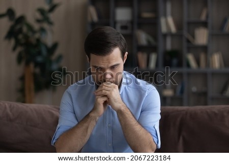 Confused unhappy young man lost in negative thoughts, considering personal problem solution, suffering from depression, feeling insecure or nervous waiting for important news, sitting on sofa at home. Royalty-Free Stock Photo #2067223184