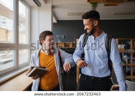 Young happy man with Down syndrome with his mentoring friend celebrating success indoors at school. Royalty-Free Stock Photo #2067232463