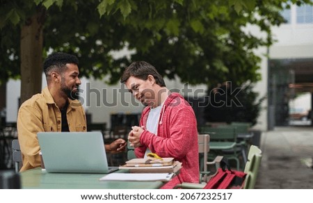 Young man with Down syndrome with his mentoring friend sitting outdoors in cafe using laptop. Royalty-Free Stock Photo #2067232517