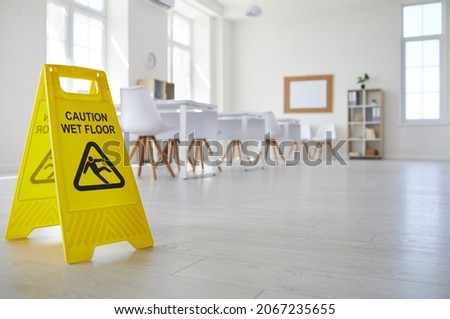 Janitorial or cleaning service plastic sign with figure that slips and falls and words Caution Wet Floor standing in clean empty classroom with white desks and chairs at school, college or university