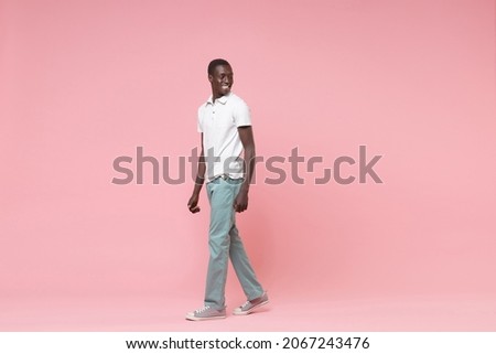 Side view of smiling young african american man guy in white polo shirt, turquoise trousers posing isolated on pastel pink wall background. People lifestyle concept. Mock up copy space. Looking aside Royalty-Free Stock Photo #2067243476