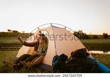 White couple taking selfie on cellphone and sitting in tent during camping together