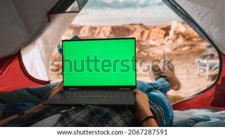 POV of a Tourist Using a Laptop Computer with Green Screen Placeholder on Display. Traveller Resting in a Tent on Top of a Rocky Mountain. Adventurous Hiker Living in Nature in Great Outdoors. Royalty-Free Stock Photo #2067287591