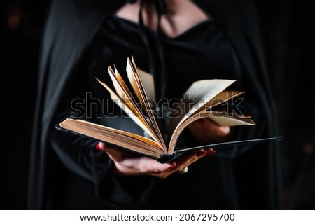 Witch in black costume reading black magic book. Halloween, spooky horror concept. Wizard outfit. Amazing hands portrait.