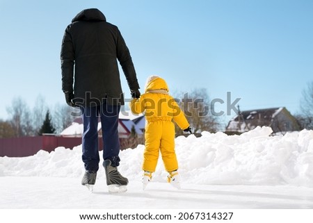 A father teaches his daughter to skate at home on his own ice rink, holding her hand on a sunny frosty winter day. A girl in a yellow jumpsuit trains skating with a personal trainer. Skating season