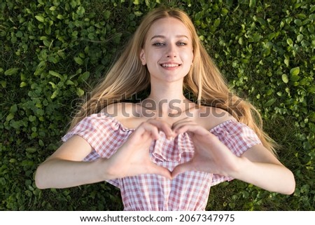 Portrait of a young and attractive Caucasian blonde girl. The girl lying on the grass in the meadow and showing a gesture of love.