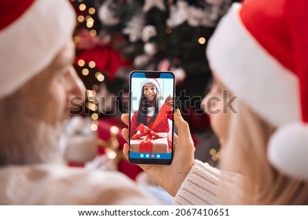 Happy senior grandparents couple in santa hats greeting grandchild opening Christmas gift on family cell phone video call, online virtual party meeting videocall on smartphone at home. Over shoulder