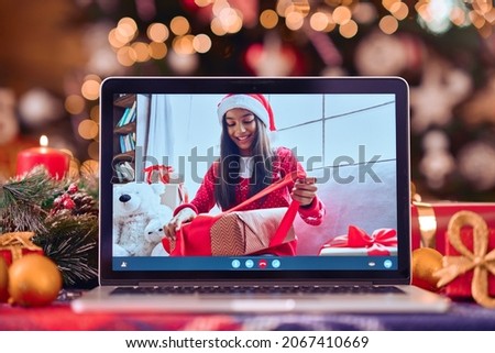 Teen girl in Santa hat opening gift box in remote chat on laptop computer screen Merry Christmas table background. Xmas online virtual party celebration, Happy New Year present unpacking videocall.