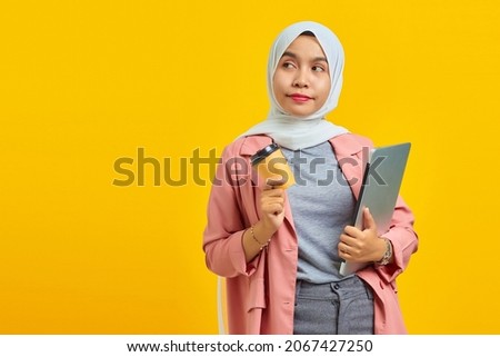 Portrait of cheerful young Asian woman holding laptop and coffee isolated over yellow background