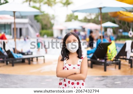 Asian child or kid girl wearing white face mask for close mouth nose and flu sick to protect coronavirus covid-19 and standing arms crossed with smile on lot of people at swimming pool or waterpark