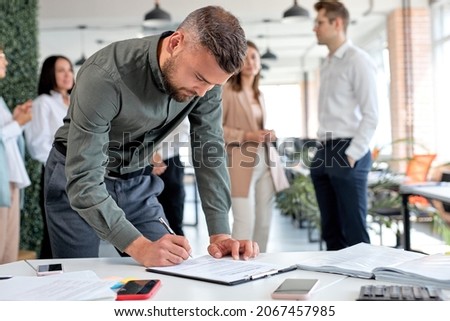 Young confident business man signing financial contract, put write signature on legal corporate paper fill document form buy insurance loan, making business agreement, colleagues in the background
