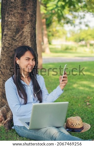 Asian woman sitting green garden park using phone listen music from smart phone with laptop computer. Happiness Woman smiling, laughing happy toothy smile working Freelance on laptop home office use.