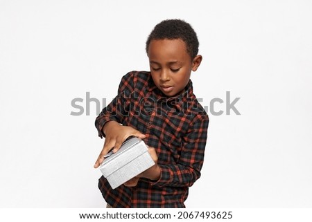 Studio portrait of handsome preschool boy with dark skin opening silver box with gift or present, holding it carefully, wearing red flannel shirt isolated on white wall with copy space for your text