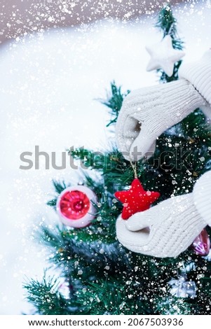 Woman’s hands with knitted white mittens gloves hanging Christmas toys on artificial fir tree branches during snowfall in winter time, copy space