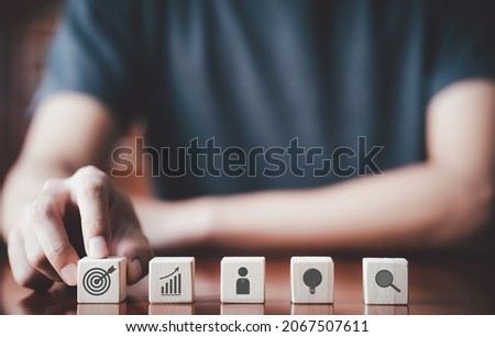 Businessman putting wooden cube with target board, chart, idea icon and on wooden table. Goals and planning for success in marketing business, achieve the objective concept. Closeup free copy space.