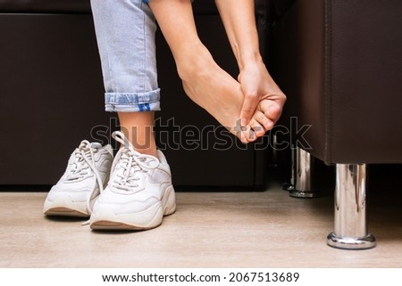 Pain in the feet from uncomfortable shoes. A woman holds a sore foot with hands sitting on a couch. Pain caused by wearing sneakers. Discomfort from walking for a long time. Orthodontics Royalty-Free Stock Photo #2067513689