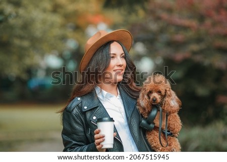 A beautiful young dark-haired woman walks in the park and hugs her poodle dog. Autumn mood. Fall.