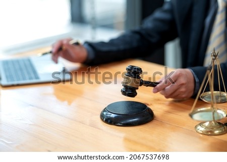 Hand of a businessman or lawyer holding a judge hammer works using a laptop keyboard the scales are placed on the office desk.