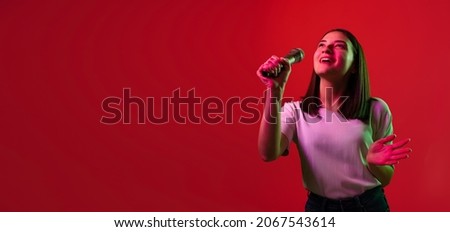 Portrait of young beautiful woman in white T-shirt singing in microphone isolated over red background in neon lights. Concept of feelings, youth, fashion, facial expression, emotions, lifestyle, ad
