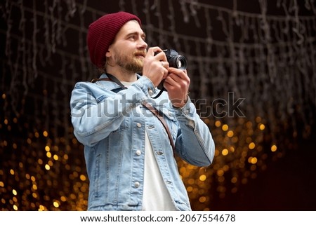 Young hipster man in denim jacket and red hat walk in city street taking photo, using retro camera, Garlands lights in the background, copy space. Photography, people lifestyle concept.