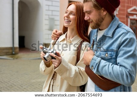 Young tourists couple taking photo of architecture on retro film camera, wearing casual outfit, standing on street of historical town. Tourist photographs sights in European town. Copyspace. Side view