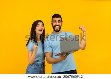 Overjoyed arab couple holding laptop and celebrating success with clenched fists, lucky winners posing with PC computer over yellow studio background, copy space