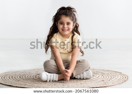 Portrait Of Cute Smiling Little Girl Sitting With Legs Crossed At Wicker Carpet At Home, Beautiful Cheerful Adorable Female Child Relaxing On Floor, Looking And Smiling At Camera, Copy Space Royalty-Free Stock Photo #2067561080