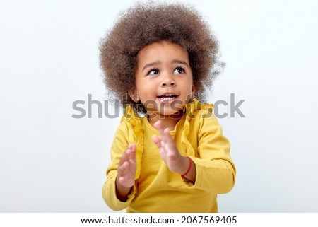 Afro fluffy child girl clapping and applauding, happy and joyful, smiling. Adorable charming kid in yellow shirt have fun isolated on white studio background, looking up. Portrait, copy space.