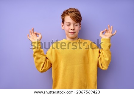 Portrait of young teenage boy keep calm, meditating, isolated on purple studio background. Kid in yellow casual shirt standing with eyes closed engaged in yoga, focused on thoughts, concentrated