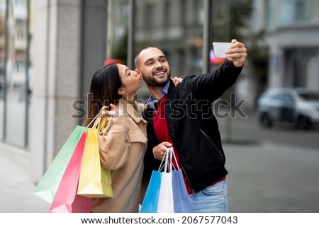 Loving interracial couple with colorful bags taking selfie on mobile phone in front of big mall. Young diverse family photographing themselves while going shopping to supermarket