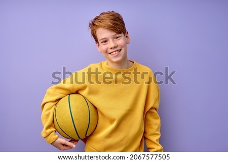Happy teenage boy in yellow shirt holding basketball ball in hands isolated in studio on purple background, portrait. pozitive excited kid looking at camera, laughing, sport, childhood concept Royalty-Free Stock Photo #2067577505