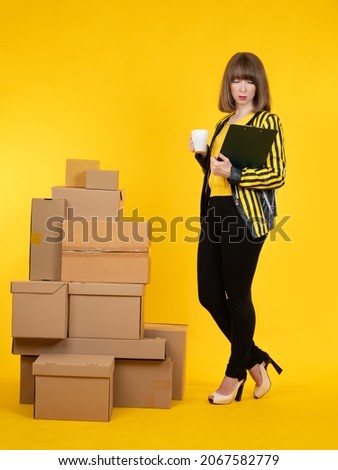 Small business owner woman. Business owner on a yellow background. Concept - she prepares goods for shipment. Corton boxes as a symbol of small business. Business woman holds a mug and clipboard.