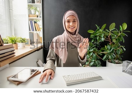 Happy young Muslim female greeting someone on computer screen by waving hand at the beginning of online lesson