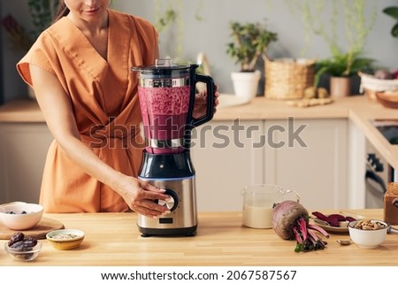 Young housewife with electric blender preparing beetroot smoothie by kitchen table with bowls containing ingredients Royalty-Free Stock Photo #2067587567