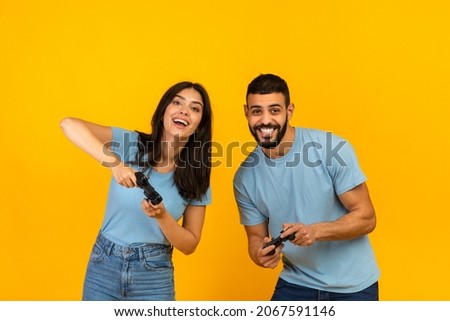 Having fun. Playful arab couple playing video games together, holding and using joysticks over yellow studio background, banner. Free time entertainment concept