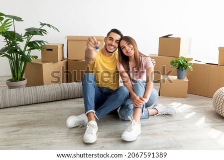 Full length portrait of loving interracial couple sitting on floor among cardboard boxes, showing house key indoors. Millennial multinational spouses relocating to new apartment together Royalty-Free Stock Photo #2067591389