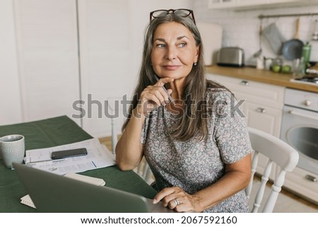 Picture of charming modern female writer holding pen looking aside creating plot line of character in new novel, sitting in kitchen in front of laptop, phone and papers. Creativity, home office