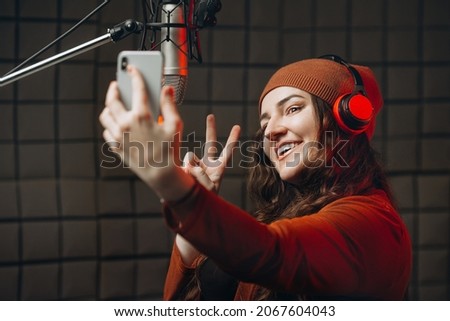 Attractive beautiful woman blogger using smartphone for selfie and recording video vlog for her subscribers at sound recording studio near a professional microphone