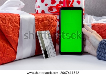 Female hands in a sweater hold a smartphone with a green vertical screen and a payment card near the gift boxes on the table. The concept of holiday shopping on the Internet . Chroma key.