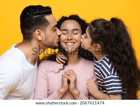Happy Mother's Day Concept. Husband And Little Daughter Kissing Happy Arab Woman In Both Cheeks, Young Middle Eastern Family Of Three Bonding Together Over Yellow Studio Background, Closeup Shot