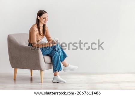 Joyful young lady sitting in armchair with modern smartphone, working remotely or video chatting against white studio wall, copy space. Millennial woman watching movie, having online meeting Royalty-Free Stock Photo #2067611486
