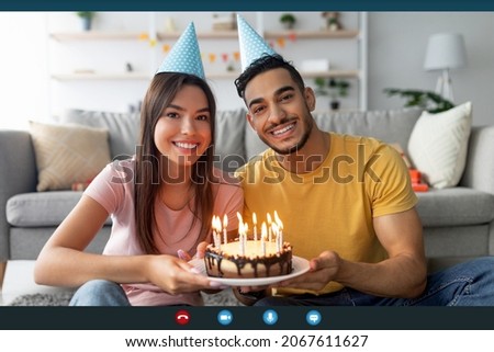 Computer screen view of happy multiracial couple in party hats showing birthday cake with candles at webcam, celebrating b-day online, making video call to family or friends on holiday, indoors