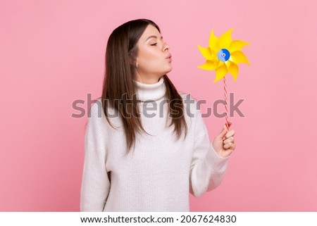 Portrait of carefree brunette woman blowing at paper windmill, playing with pinwheel toy on stick, wearing white casual style sweater. Indoor studio shot isolated on pink background. Royalty-Free Stock Photo #2067624830
