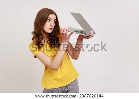 Woman with curly hair in urban style yellow T-shirt peeking out half-closed laptop with sly suspicious look, spying and knowing secret information. Indoor studio shot isolated on gray background.