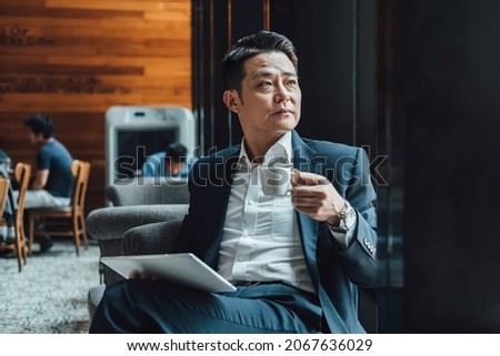 Handsome Asian Businessman Using Digital Tablet in a Cafe. 
Serious business man in blue suit working on a digital tablet while sitting in a restaurant and drinking cup of coffee. Royalty-Free Stock Photo #2067636029